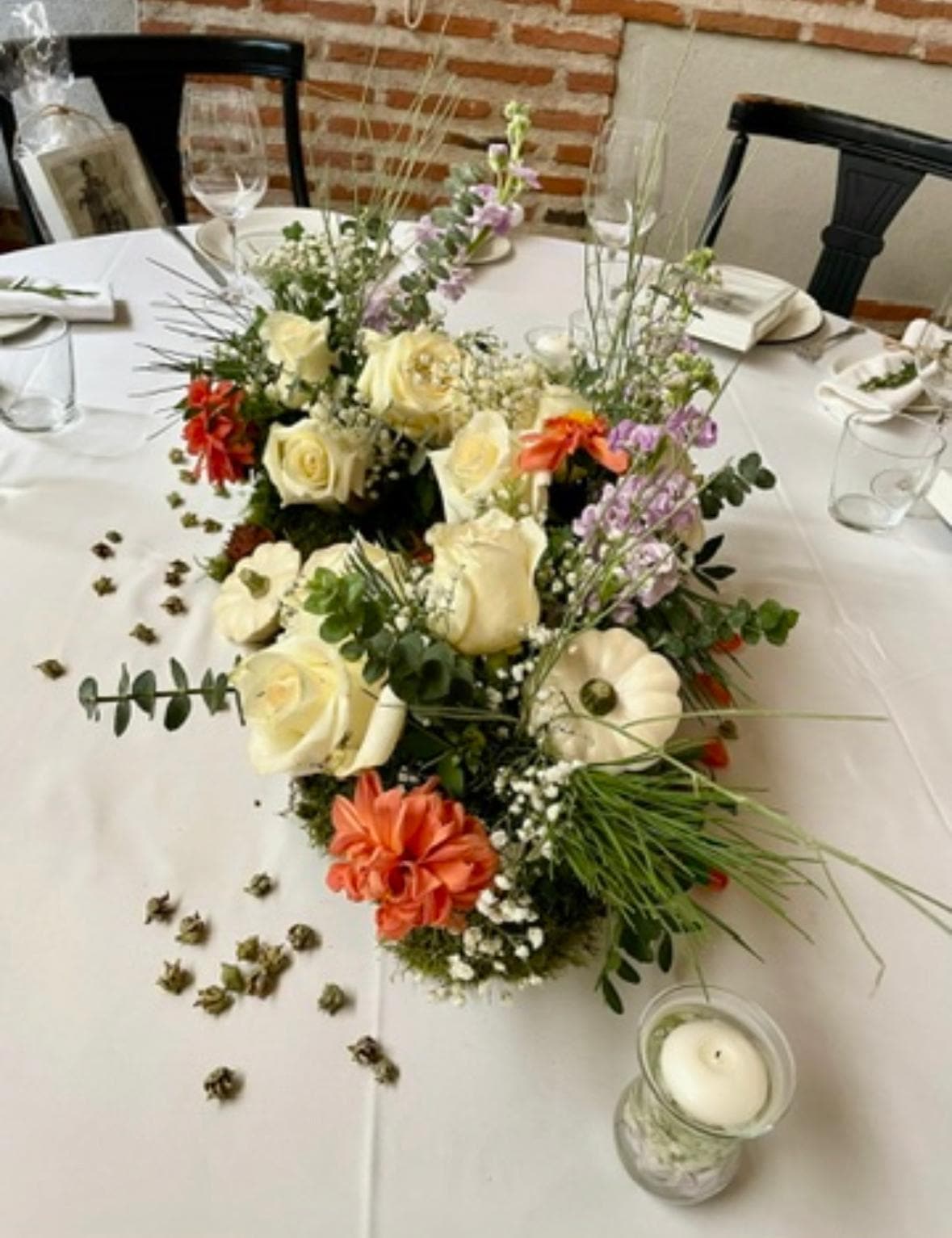 Centro de mesa con rosas blancas, dalias coral y vela junto a pared de ladrillo