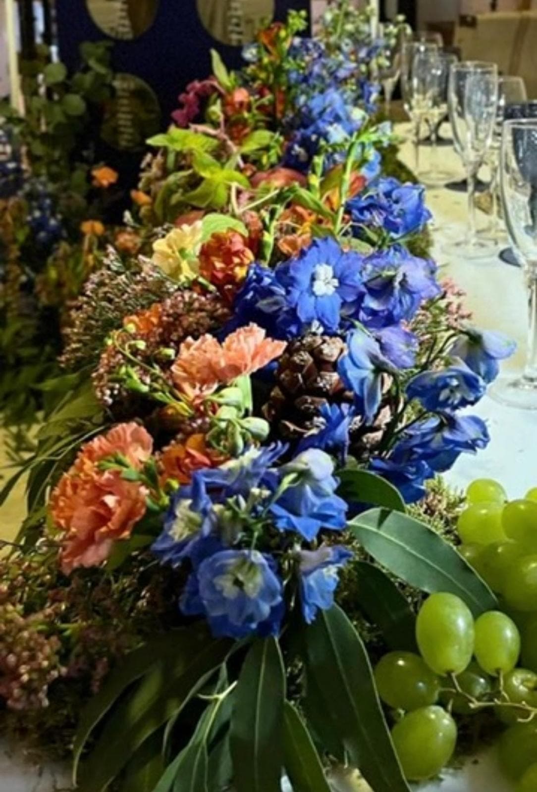 Camino de mesa con delphinium azul, claveles naranjas y uvas